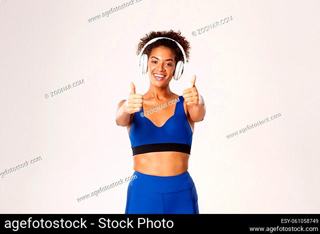 Happy african-american female athelte in blue sportswear, listening music in headphones during workout, showing thumbs-up, recommend something, white background