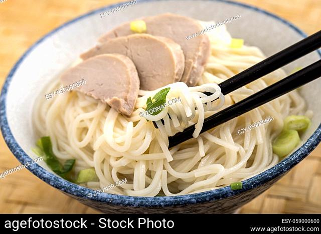A close up photo of using chopsticks to eat a bowl of noodles topped with pork slices and green onions