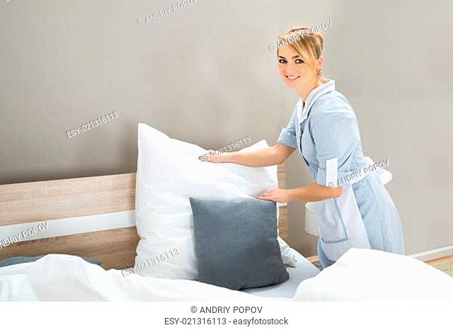 Young Female Happy Housekeeping Worker Arranging Pillows In Bedroom