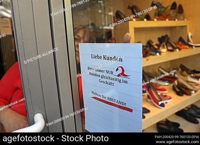 20 April 2020, North Rhine-Westphalia, Cologne: A shopkeeper hangs a sign in the shop window with the inscription ""Please only two customers in the shop at the...