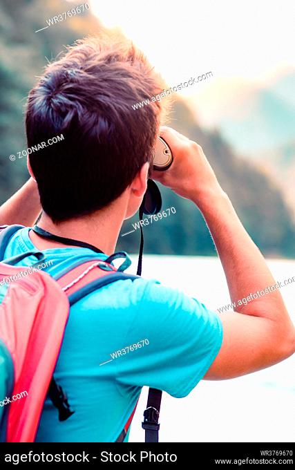Young tourist with backpack looks through a binoculars on mountains peaks, stands on a rock over a river. Boy spends a vacation in mountains