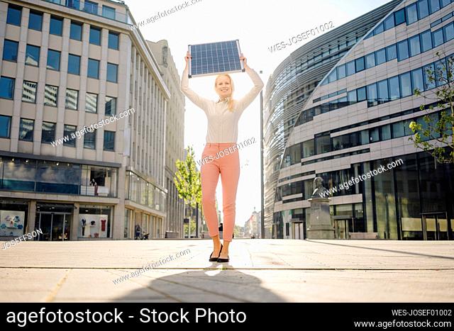 Smiling beautiful female professional holding solar panel while standing on footpath in financial district