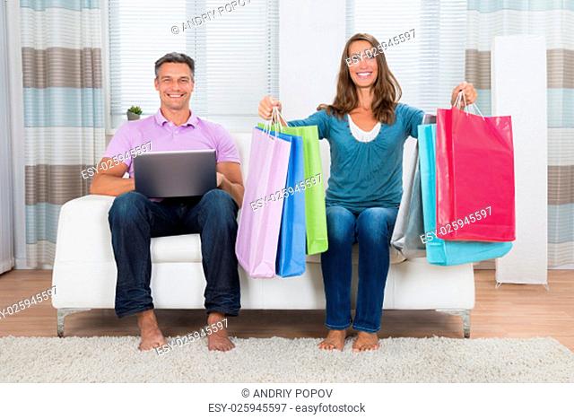 Mature Happy Couple With Shopping Bags And Laptop Sitting On Couch