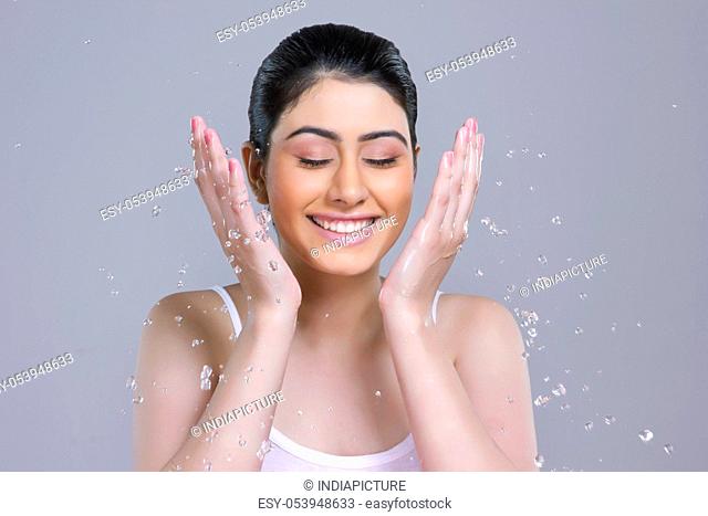 Smiling young woman washing face with water over gray background