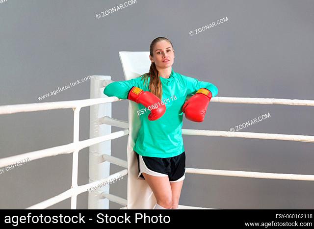 Beautiful athletic young woman with collected hair wearing in green long sleeve and red gloves standing, leaning near boxing ring and looking at camera