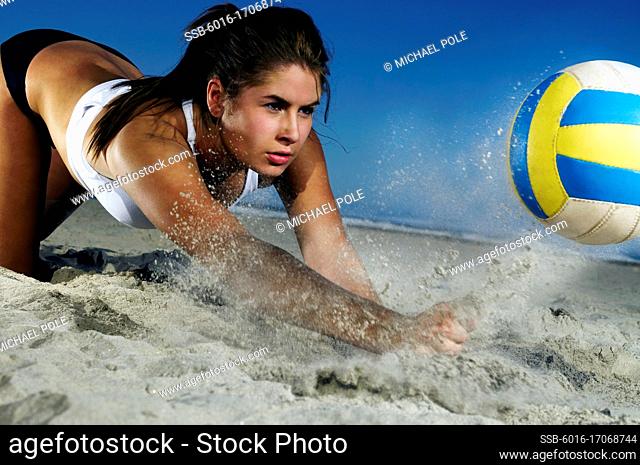 Young woman lunging onto sand to hit a volleyball upwards before it hits the ground