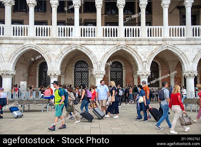 tourists with rolling suitcases passing Palazzo Ducale in the back at San Marco square in Venice, Italy