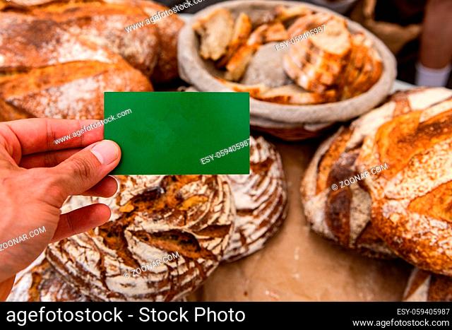 A point of view from a local baker selling rustic bread loaves at a farmer's market. Person's hand is seen holding blank green business card with copy space