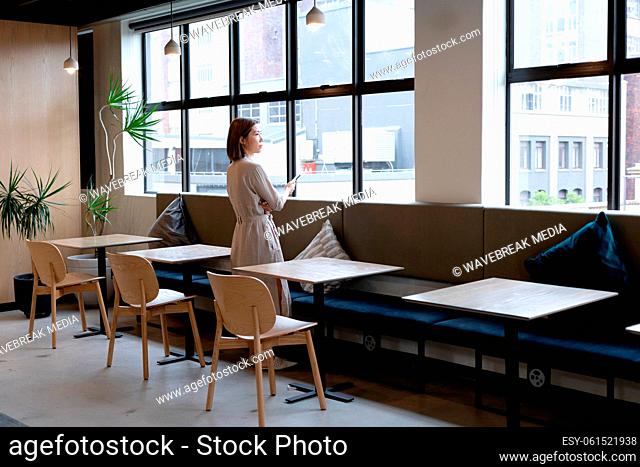 Asian woman with smartphone looking out of window in office cafeteria