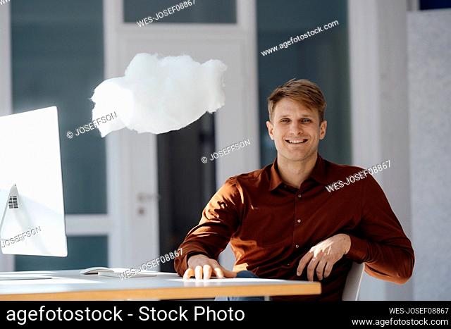 Smiling businessman with desktop PC sitting by levitating cloud network in office