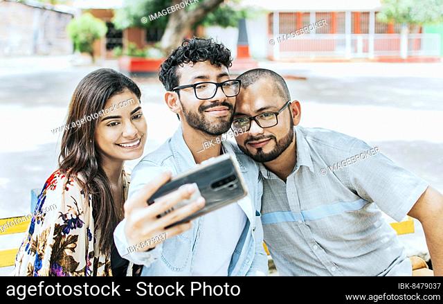 Three smiling friends sitting on a bench taking a selfie. Front view of three happy friends taking a selfie while sitting on a bench