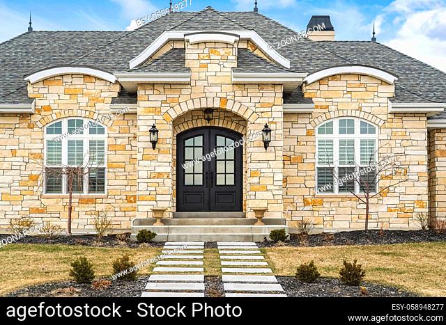 Home facade with stone brick wall double glass paned door and arched windows. Footpath on a grassy yard and doorstep lead to the arched entrance