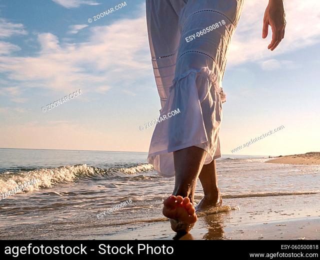 Closeup of woman wearing white skirt strolling barefooted along the beach
