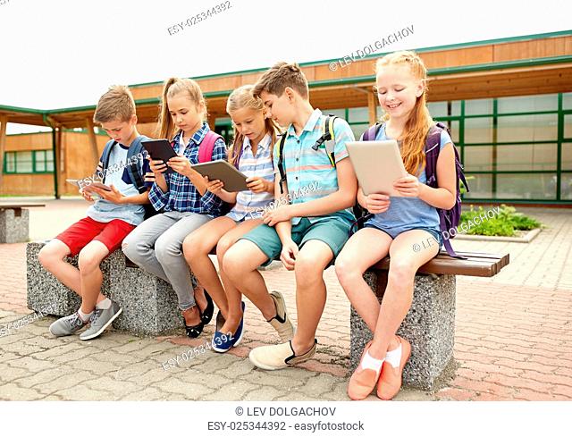 primary education, friendship, childhood, technology and people concept - group of happy elementary school students with backpacks sitting on bench and talking...