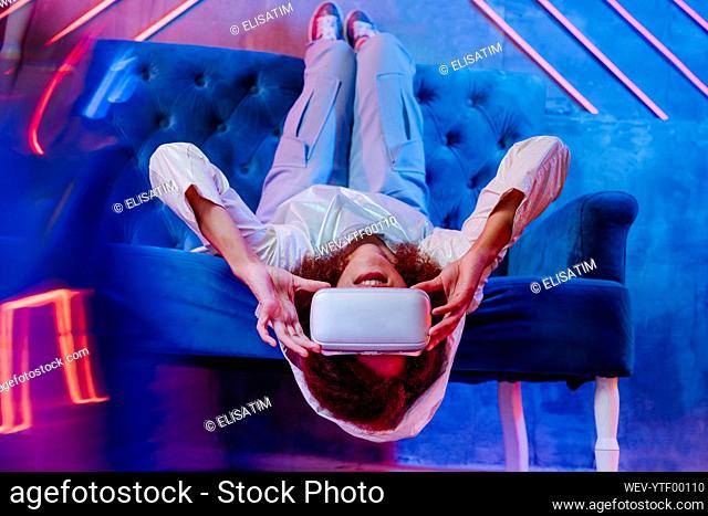 Woman with VR glasses lying upside down on sofa in front of neon light