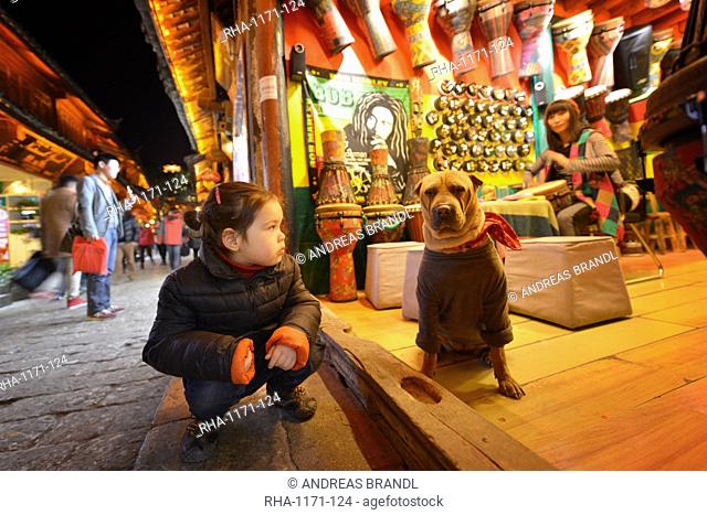 Young girl sitting next to a dog that is wearing bandana and sweater, Lijiang, Yunnan, China, Asia