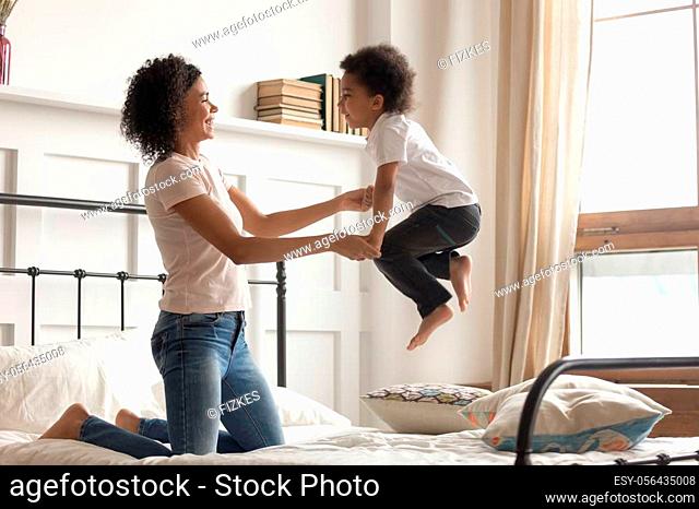 Happy african american family loving single mom holding hands of cute little kid boy laugh jumping on bed, young mixed race mother having fun playing funny...
