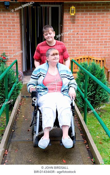 Young carer pushing his mother, who is severely asthmatic, in a wheel chair down a ramp outside their house