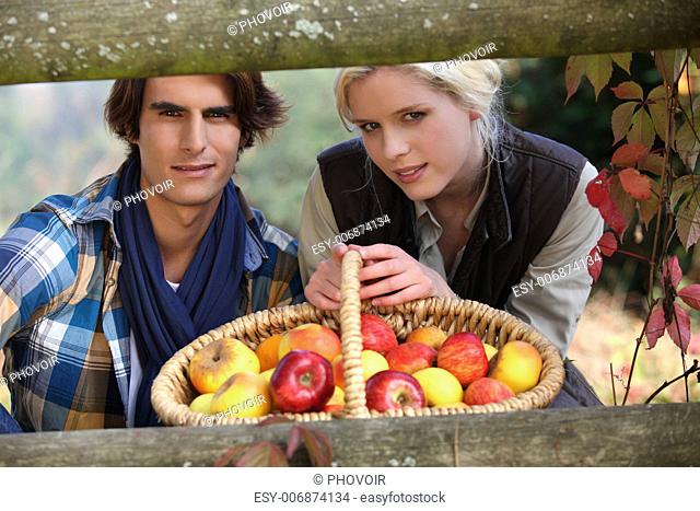 a young couple posing behind a wooden barrier with a wickerwork basket full of apples