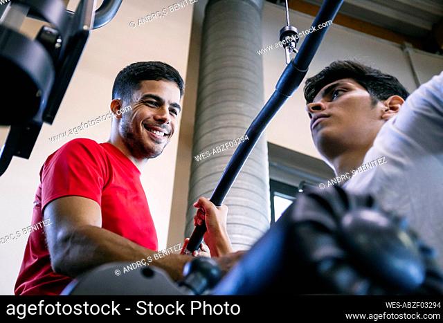Smiling fitness instructor assisting man in exercising with lateral pull-down weights at gym