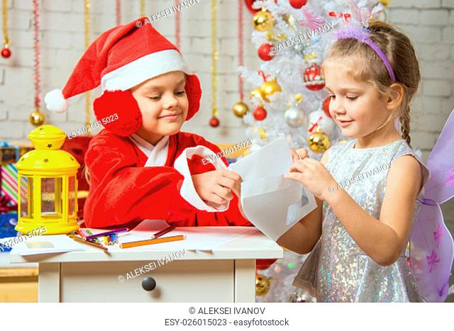 A girl dressed in a red suit Santa Claus draws pencils sitting at the table, next is a girl dressed as a fairy with the candlestick