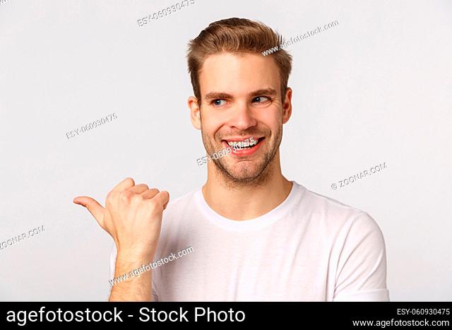 Intrigued and sly good-looking blond bearded guy with bristle in white t-shirt, peeking and pointing left with excited, thrilled smile, standing enthusiastic