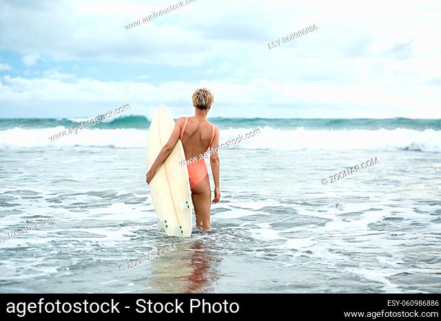 Pretty blonde girl with short hairstyle stands backsides in the waves on the beach on the background of the sea and the cloudy sky