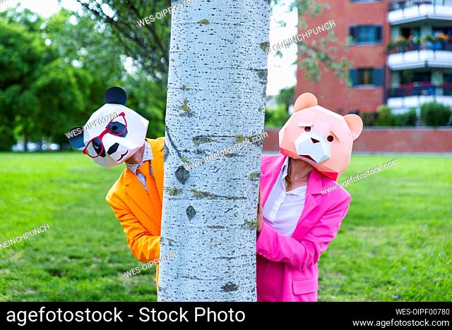 Man and woman wearing vibrant suits and animal masks hiding together behind birch tree