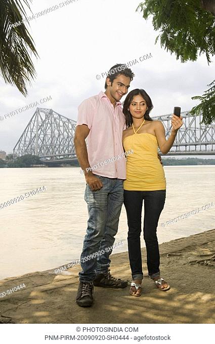Couple taking a picture of themselves with a bridge in the background, Howrah Bridge, Hooghly River, Kolkata, West Bengal, India