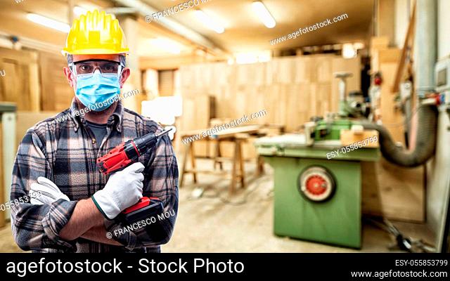 Carpenter worker in the carpentry workshop, wears helmet, goggles, leather gloves and surgical mask to prevent coronavirus infection