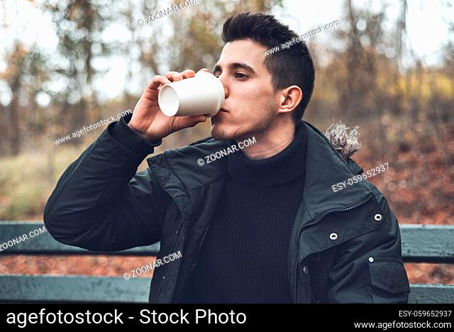 Young man sitted in a bench holding disposable coffee cup in the park in autumn season