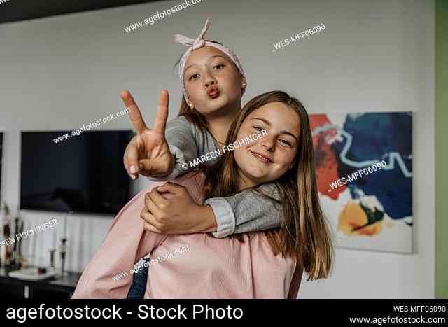 Smiling girl piggybacking friend showing victory sign while standing at home