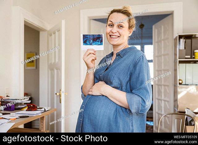 Portrait of happy pregnant woman showing ultrasound image at home