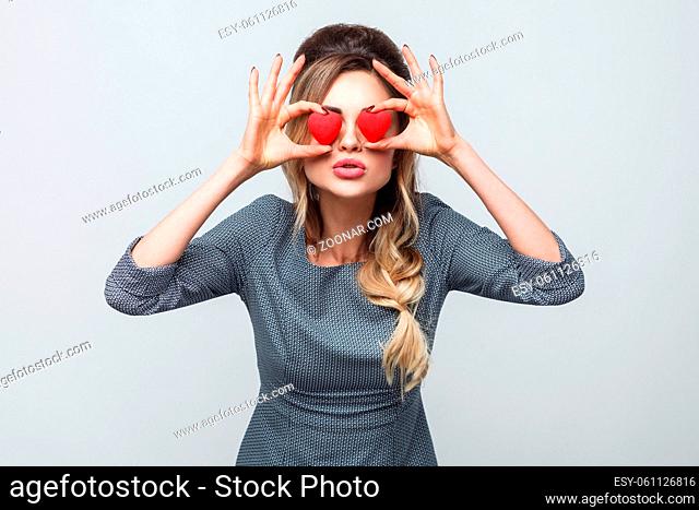 Loving eyes. Beautiful romantic caucasian young woman holding two valentine hearts in front of her eyes like glasses while standing against grey background