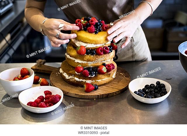 a cook working in a commercial kitchen arranging fresh fruit over a layered cake with fresh cream