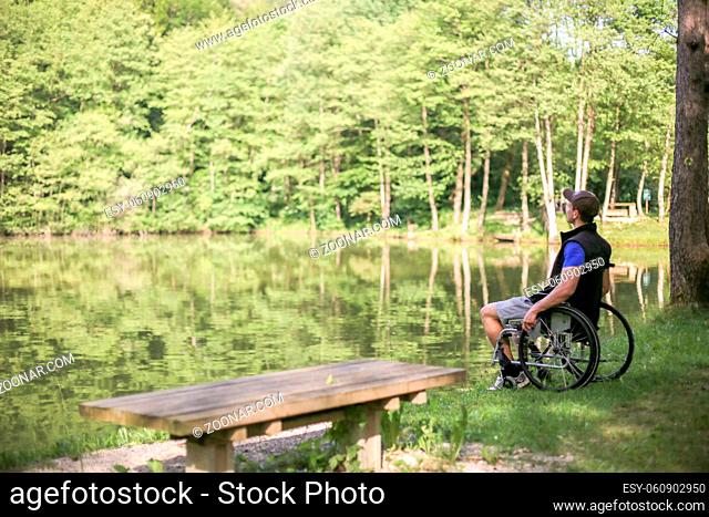 Happy and young disabled or handicapped man sitting on a wheelchair looking at beautiful lake in nature