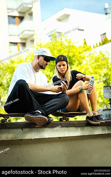 boy and girl on a skateboard are sitting and watching something on the mobile phone on a summer day