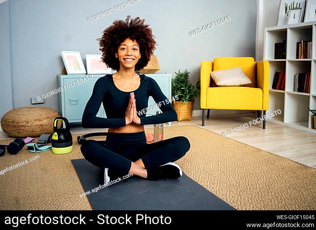 Happy young woman sitting with hands clasped on exercise mat in living room at home