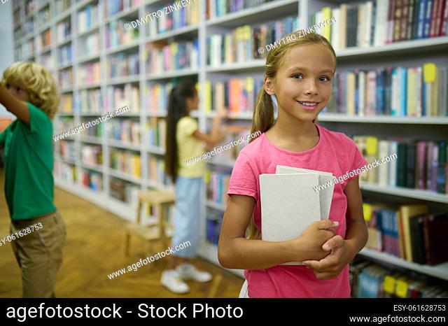 Joyous pretty teenage girl with a pile of book in her hands and her friends standing among the bookshelves