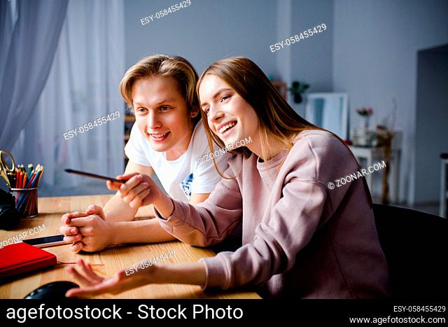 Two young belovers sit together at the table. A smiling girl shows pictures on PC his boyfriend