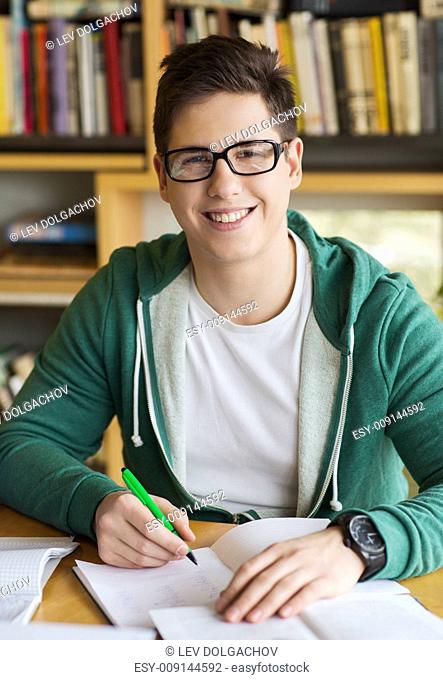 people, knowledge, education and school concept - happy student in eyeglasses with book writing to notebook in library
