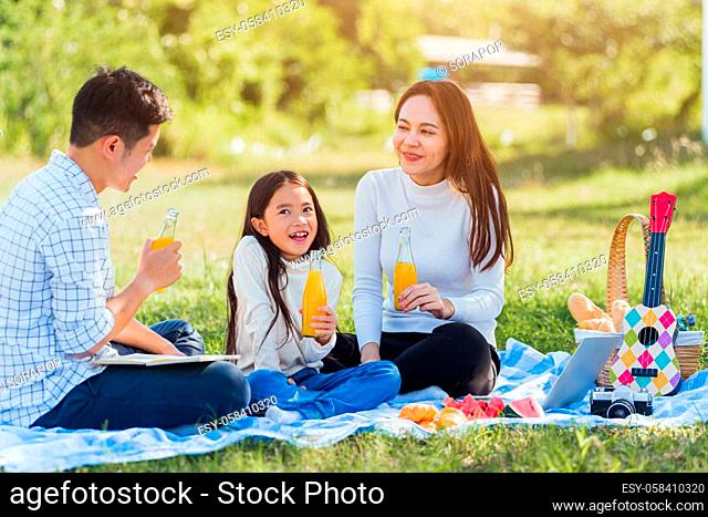 Happy Asian young family father, mother and child little girl having fun and enjoying outdoor sitting on picnic blanket drinking orange juice from glass bottle