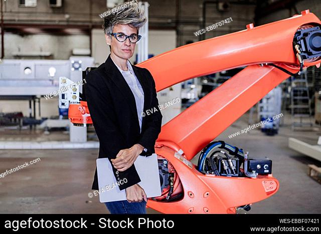 Confident mature businesswoman with laptop standing by robotic arm in industry