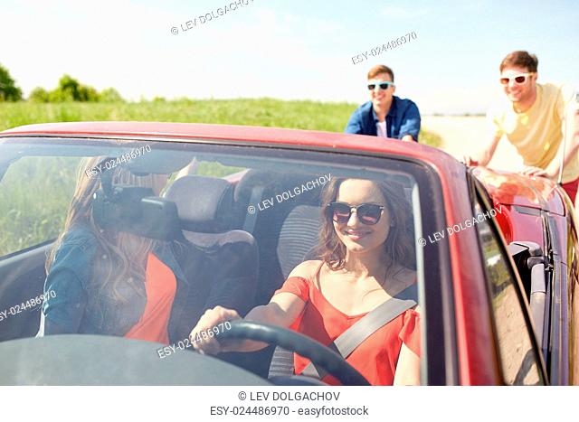 leisure, road trip, travel and people concept - happy friends pushing broken cabriolet car along country road