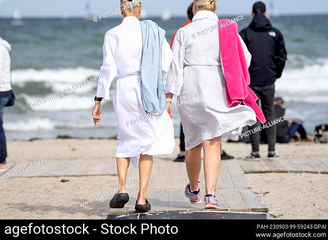 03 September 2023, Mecklenburg-Western Pomerania, Rostock Warnemünde: Two women in bathrobes walk on a beach access towards the water Baltic Sea beach of...