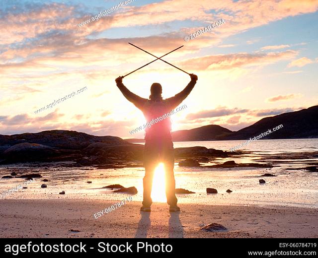Man hiker with a backpack and trekking pole standing at rocky shore and looking on the evening horizon. Sun flare in the lens