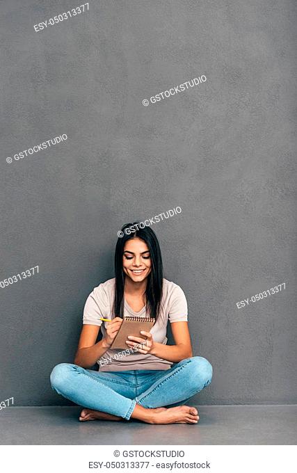 Making some notes. Attractive young woman in casual wear making notes in her notebook and smiling while sitting barefoot and against grey background