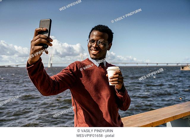 Young man taking smartphone selfie at the sea, holding take out coffee
