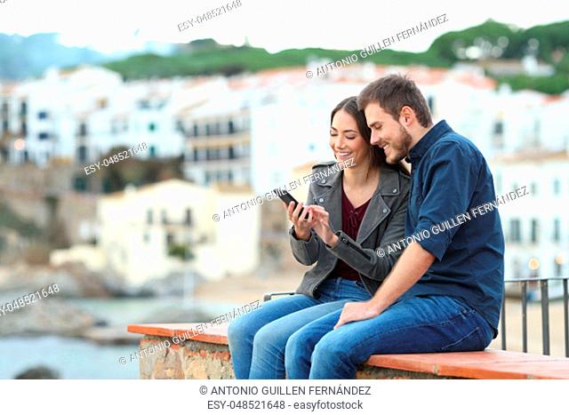 Happy couple on a ledge checking smart phone with a town in the background
