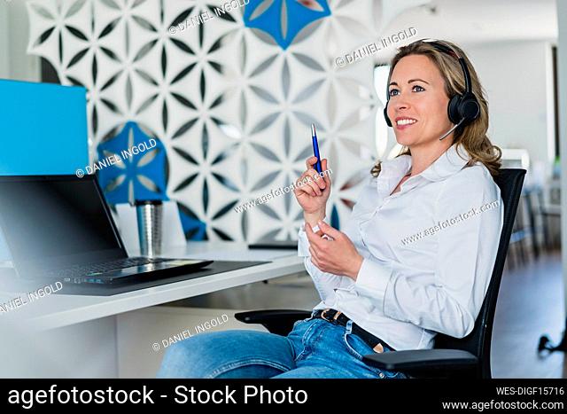 Smiling female customer service representative looking away while talking through headphones at desk